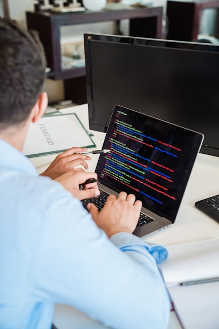 A person wearing a light blue shirt is seated at a desk, working on a laptop that displays lines of colorful code on the screen. The desk has a black computer monitor positioned behind the laptop, sho